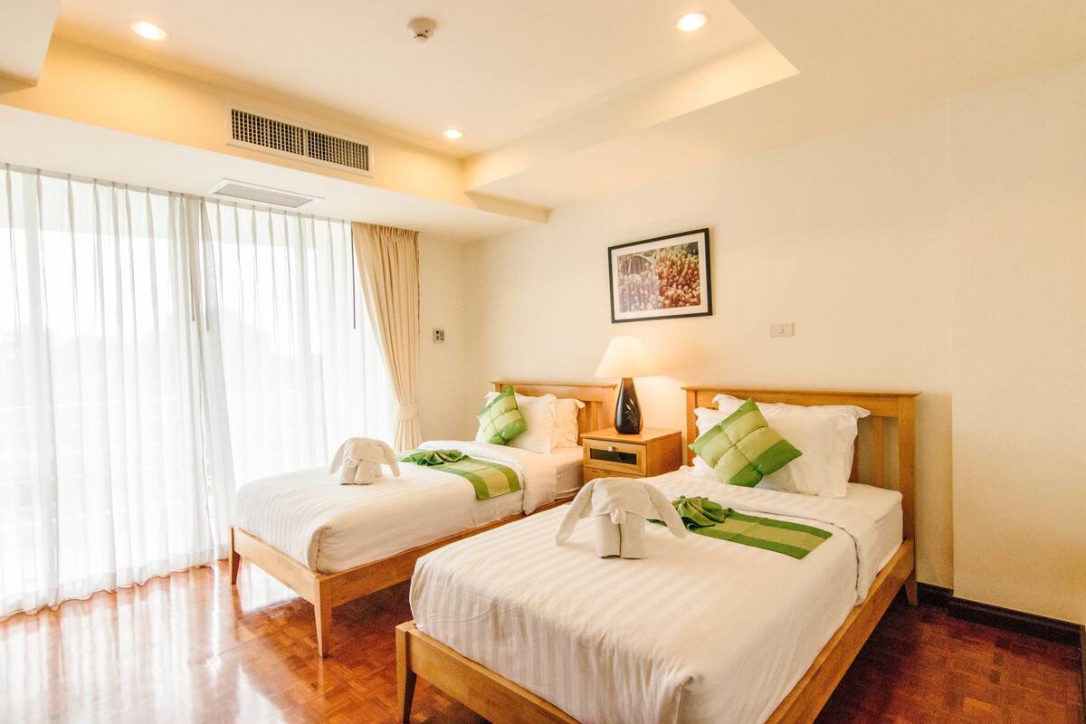 A hotel room with two beds, a large window with curtains, a wall-mounted picture, and a bedside table with a lamp.