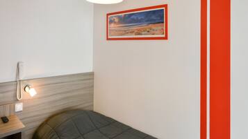 A single bed with a grey quilt, a bedside table with a lamp, a phone, and a framed picture on the wall.