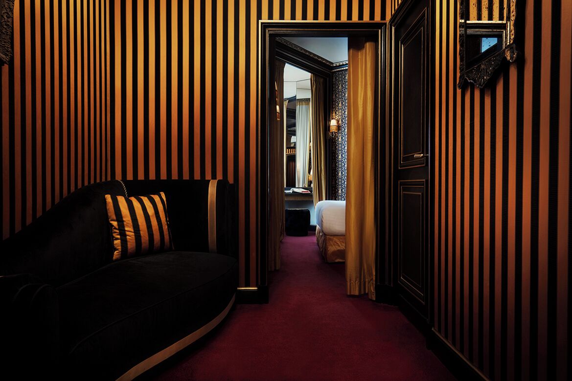A narrow hallway with striped wallpaper, a black sofa with striped cushions, and a bed in the distance.