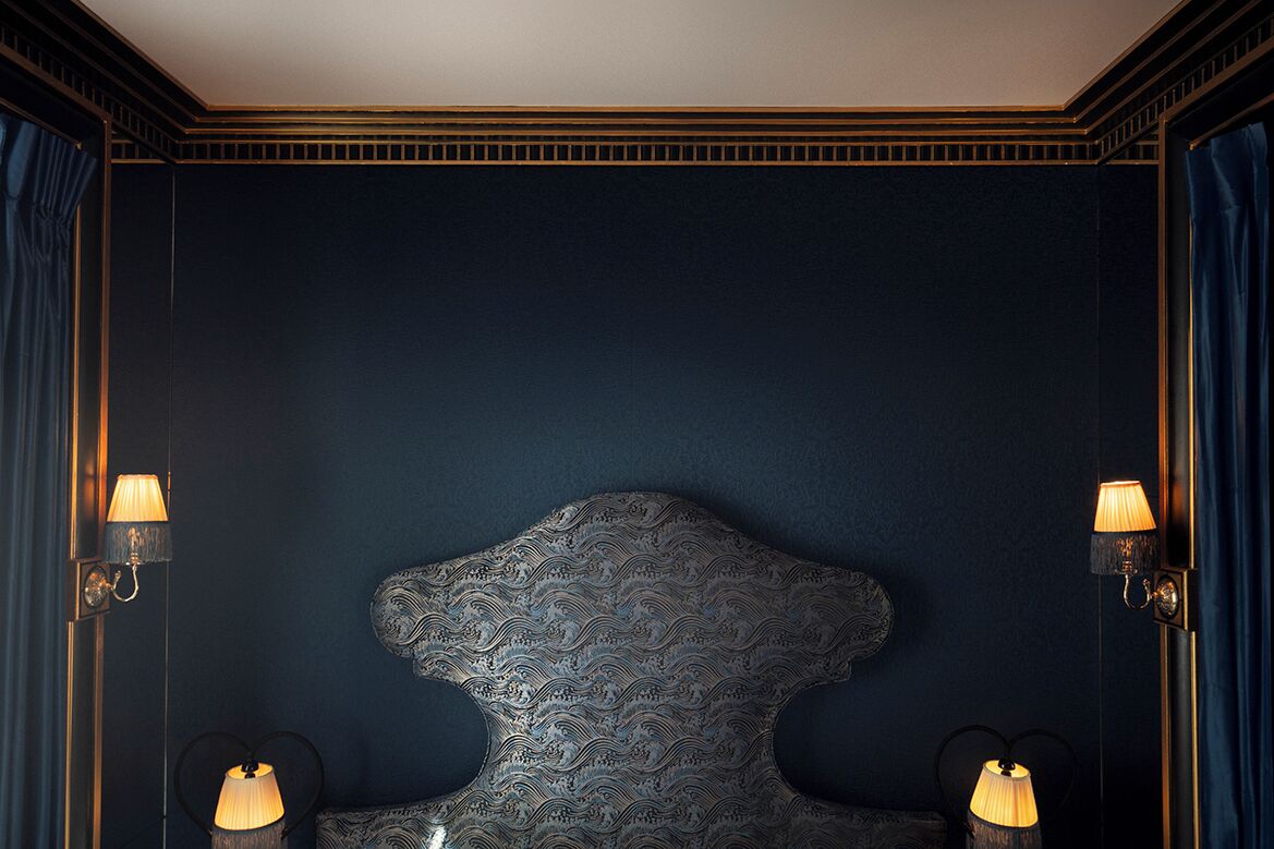 Une chambre à l’ambiance sombre, dotée d’un grand et somptueux panneau de tête de lit, de lampes fixées au mur et d’un rideau.