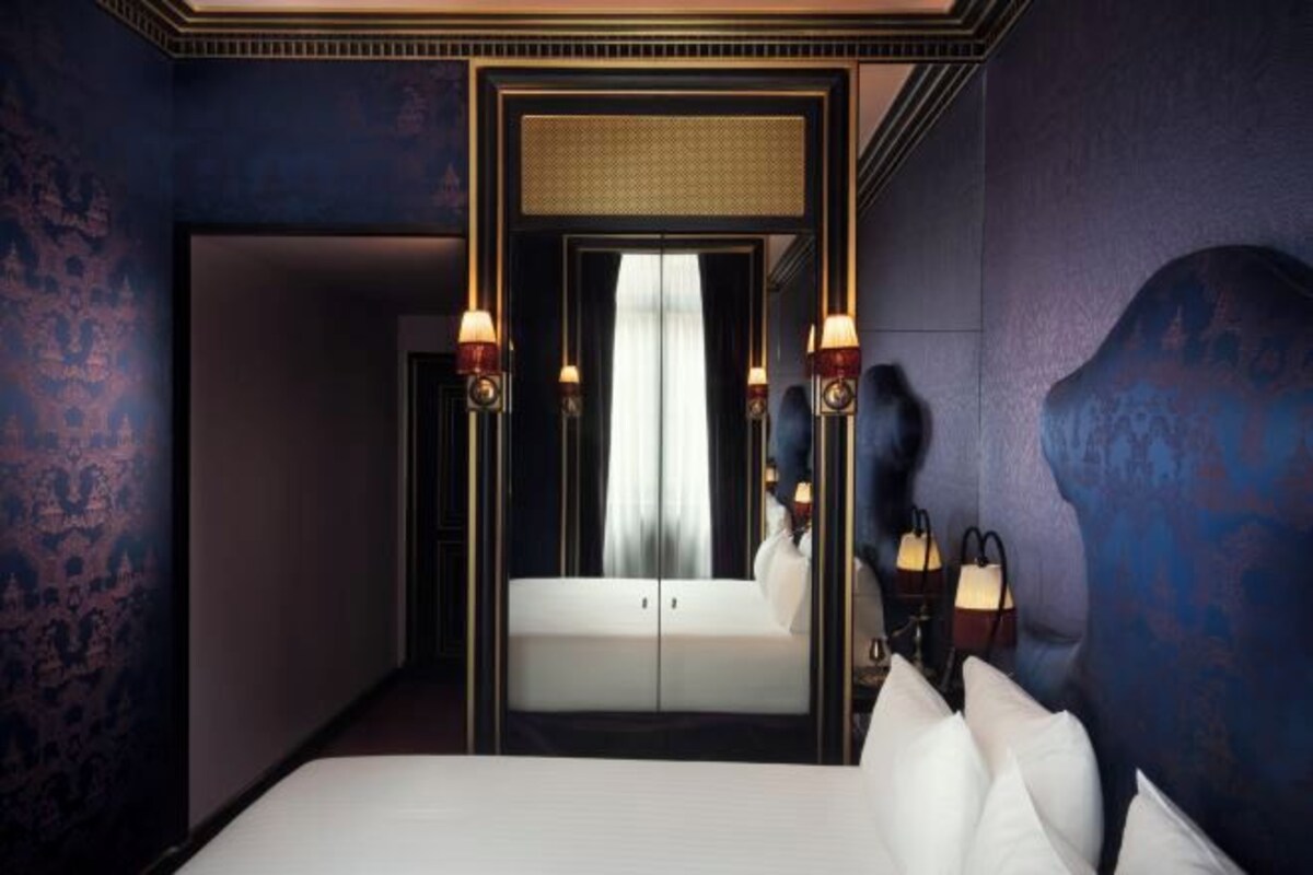 A hotel room with a large bed, a mirror, two wall-mounted lamps, and a view of a white sofa through a door.