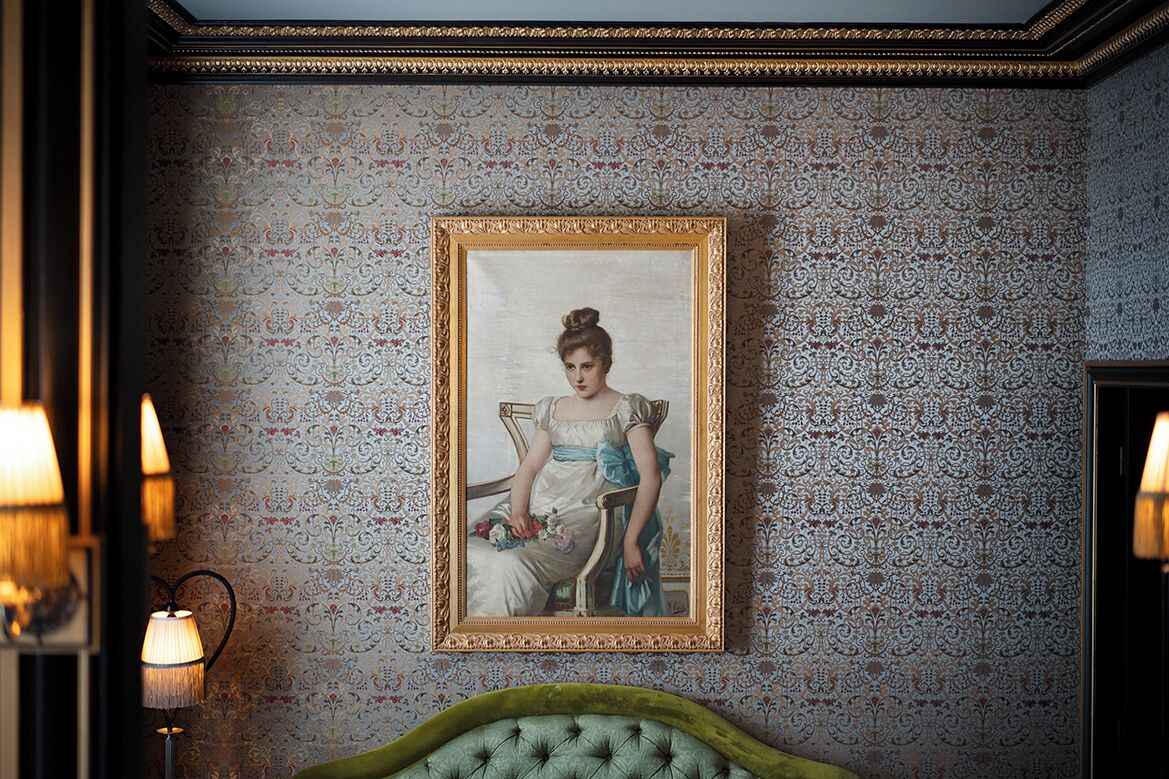 A room with a patterned wallpaper, a framed portrait on the wall, and a green upholstered headboard.