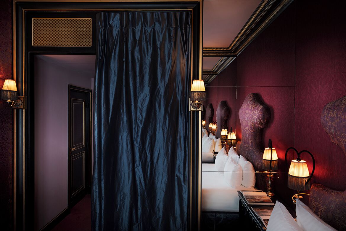 Une chambre d’hôtel avec des rideaux bleu foncé, un lit recouvert de draps blancs et des lampes fixées au mur.