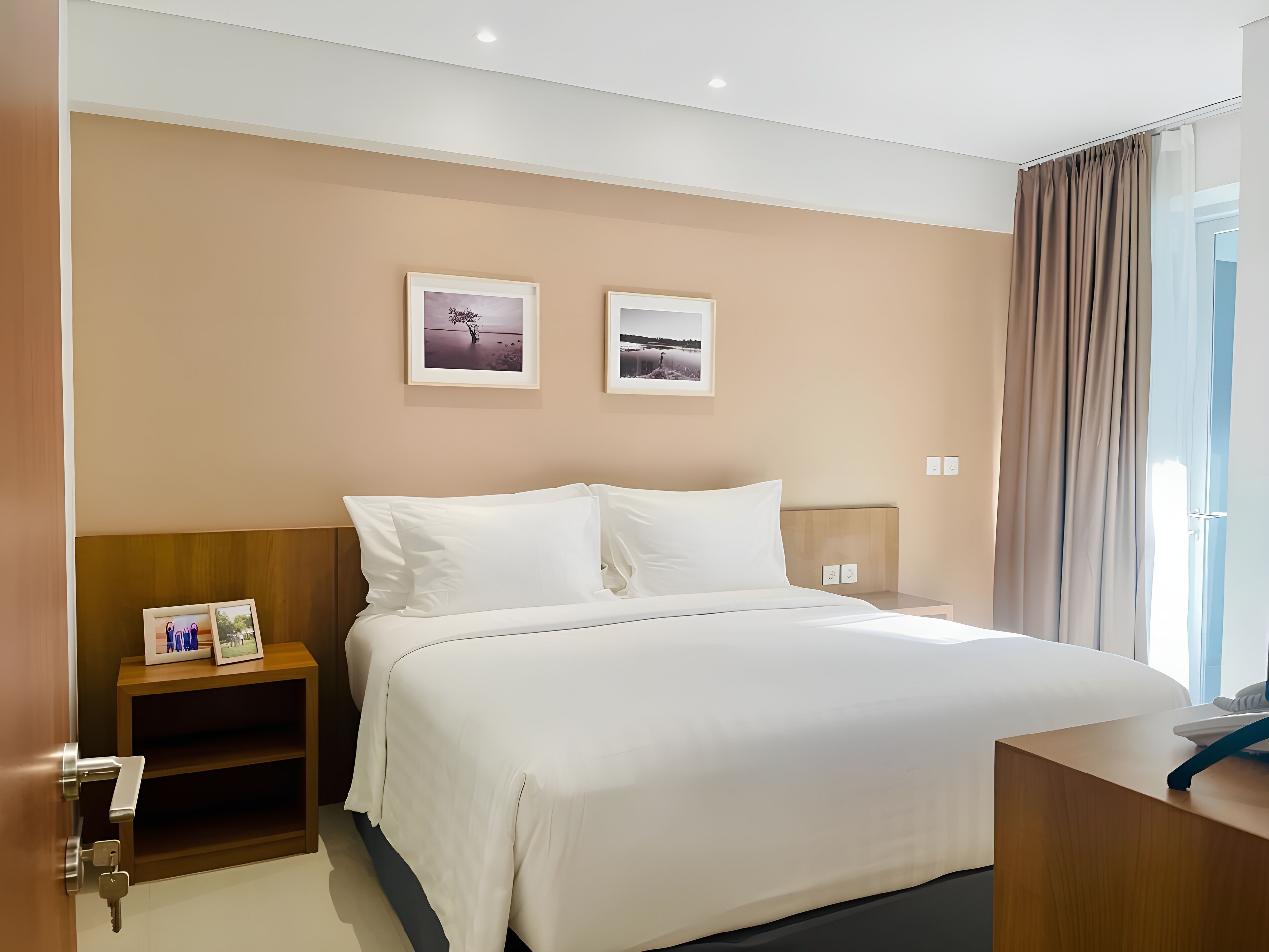 Una camera d'albergo con un letto grande, un comodino in legno, due quadri appesi alla parete e vista all'esterno attraverso una finestra con tende.