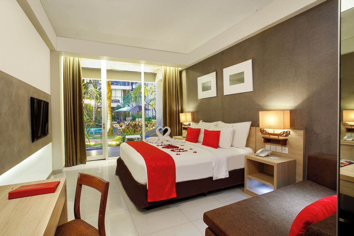 Habitación de hotel con cama, escritorio, silla, mesita de noche con lámpara y vistas al exterior.