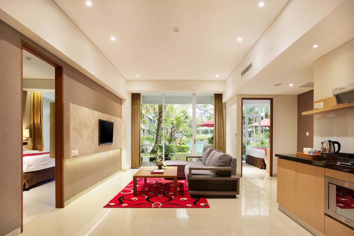 A modern hotel room with a sofa, a coffee table, a TV mounted on the wall, and a kitchen area.