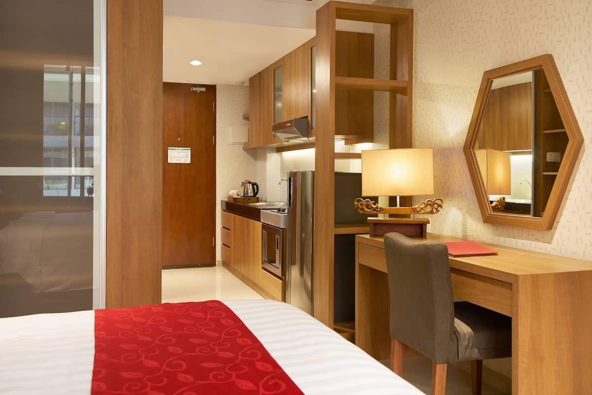 A hotel room with a bed, a desk with a lamp, a mirror, a kitchenette, and a wooden wardrobe.