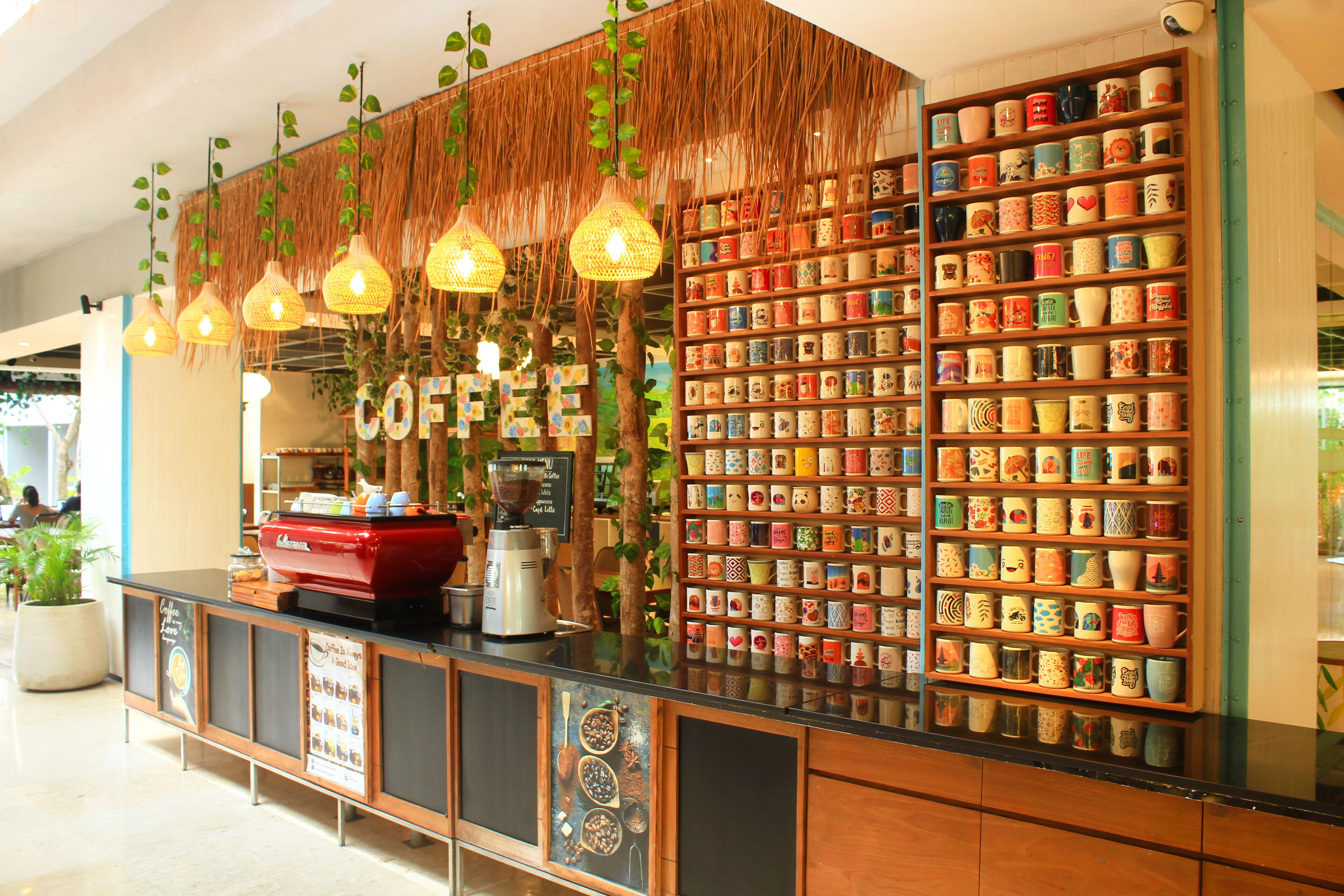 A coffee shop counter with a coffee machine, a display of colorful mugs, and hanging lights.