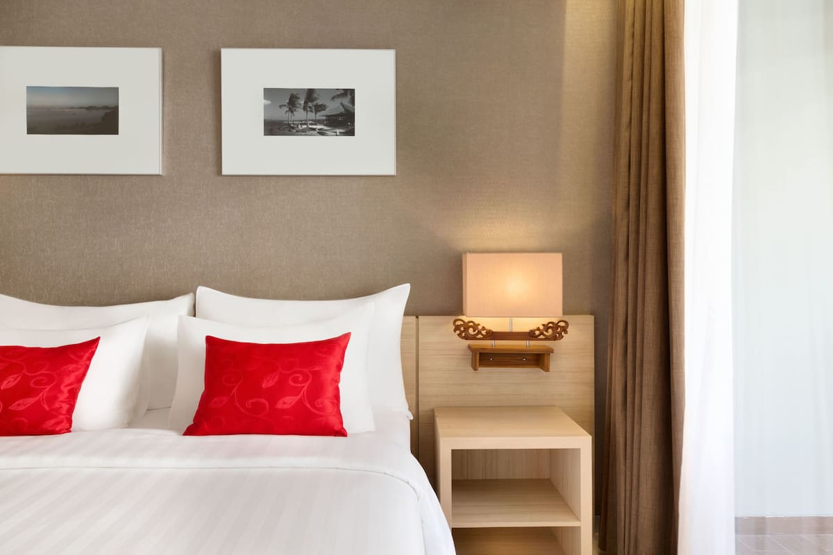 A hotel room with a bed, two red pillows, a nightstand with a lamp, and two framed pictures on the wall.