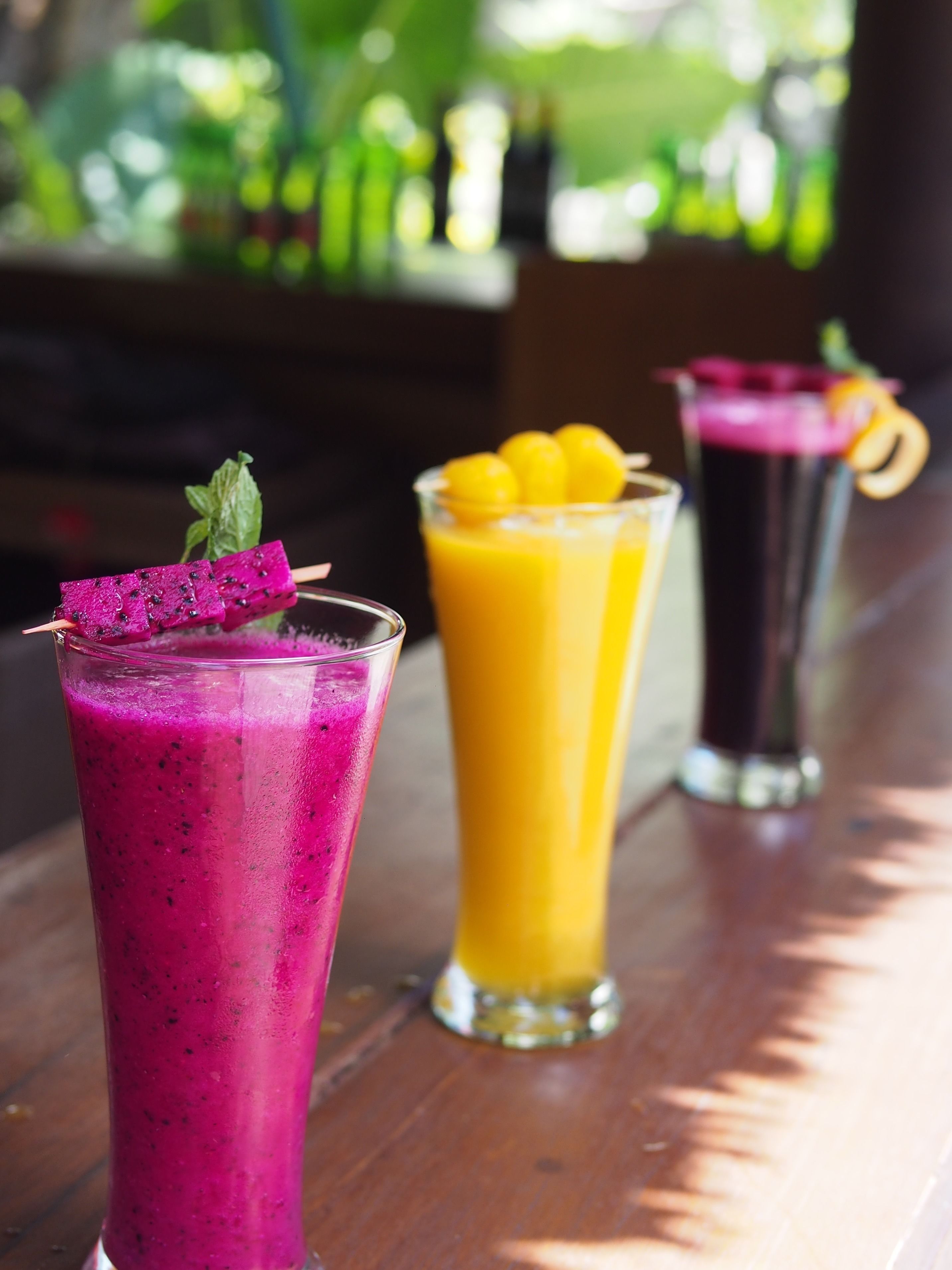 Three tall glasses of colorful smoothies on a wooden table with a blurred outdoor background.