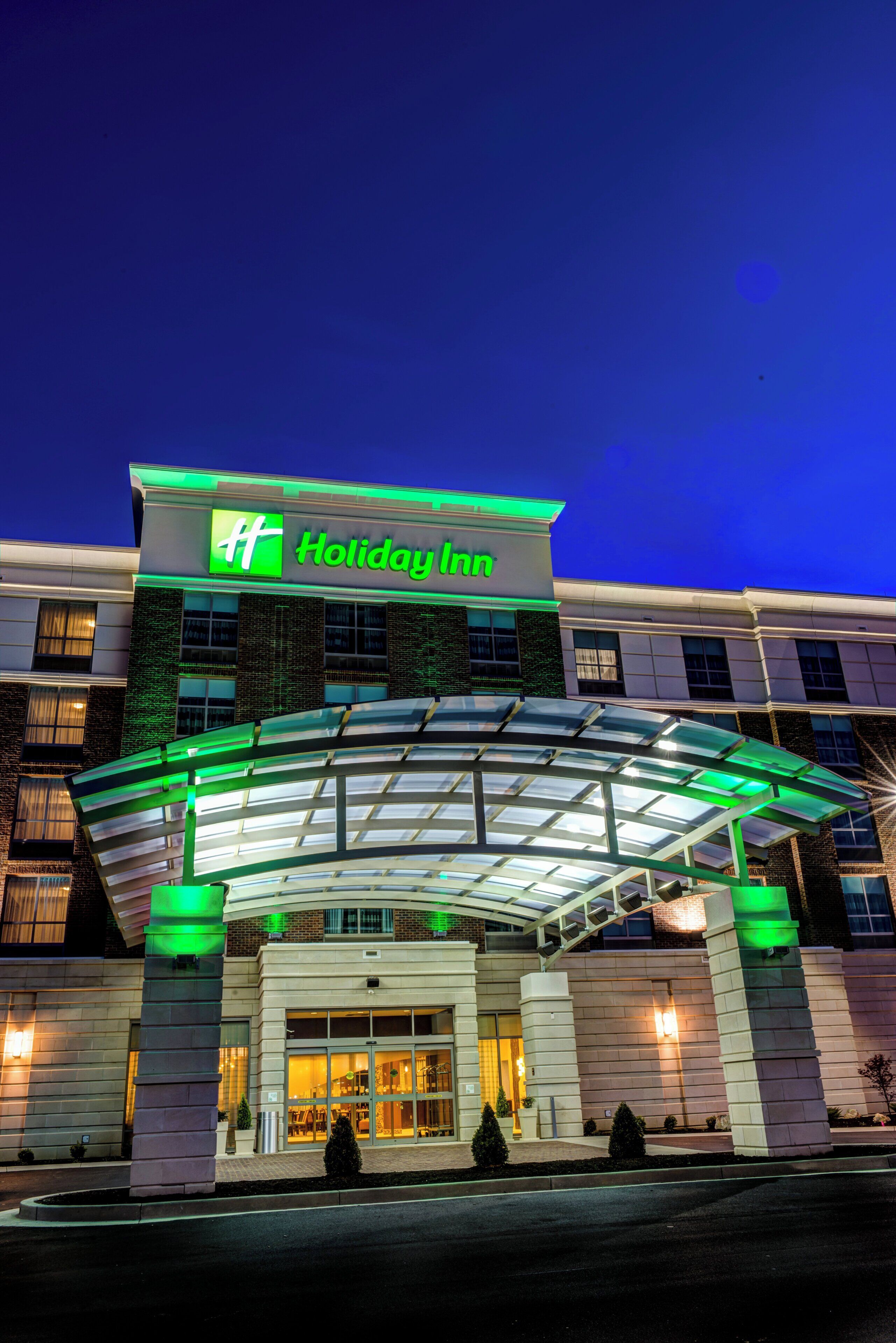 A Holiday Inn hotel with a modern design, featuring a prominent green and white logo, a covered entrance, and a brick facade.