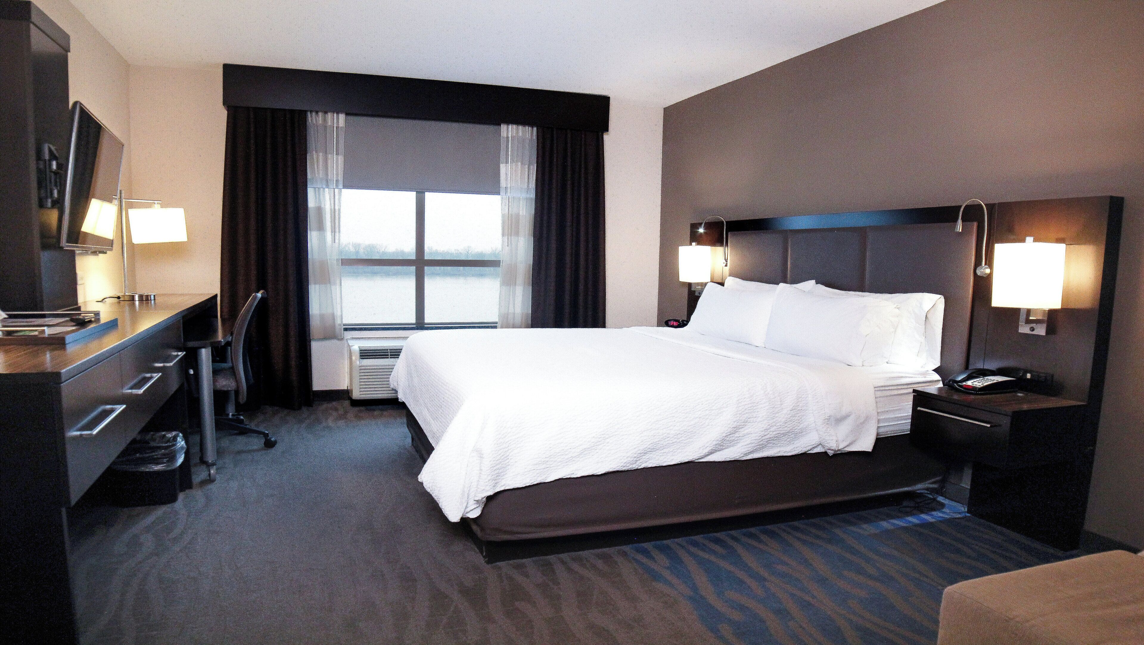 Une chambre d’hôtel avec un grand lit, des lampes de chevet, un bureau avec une lampe, une chaise et une vue sur une étendue d’eau par la fenêtre.