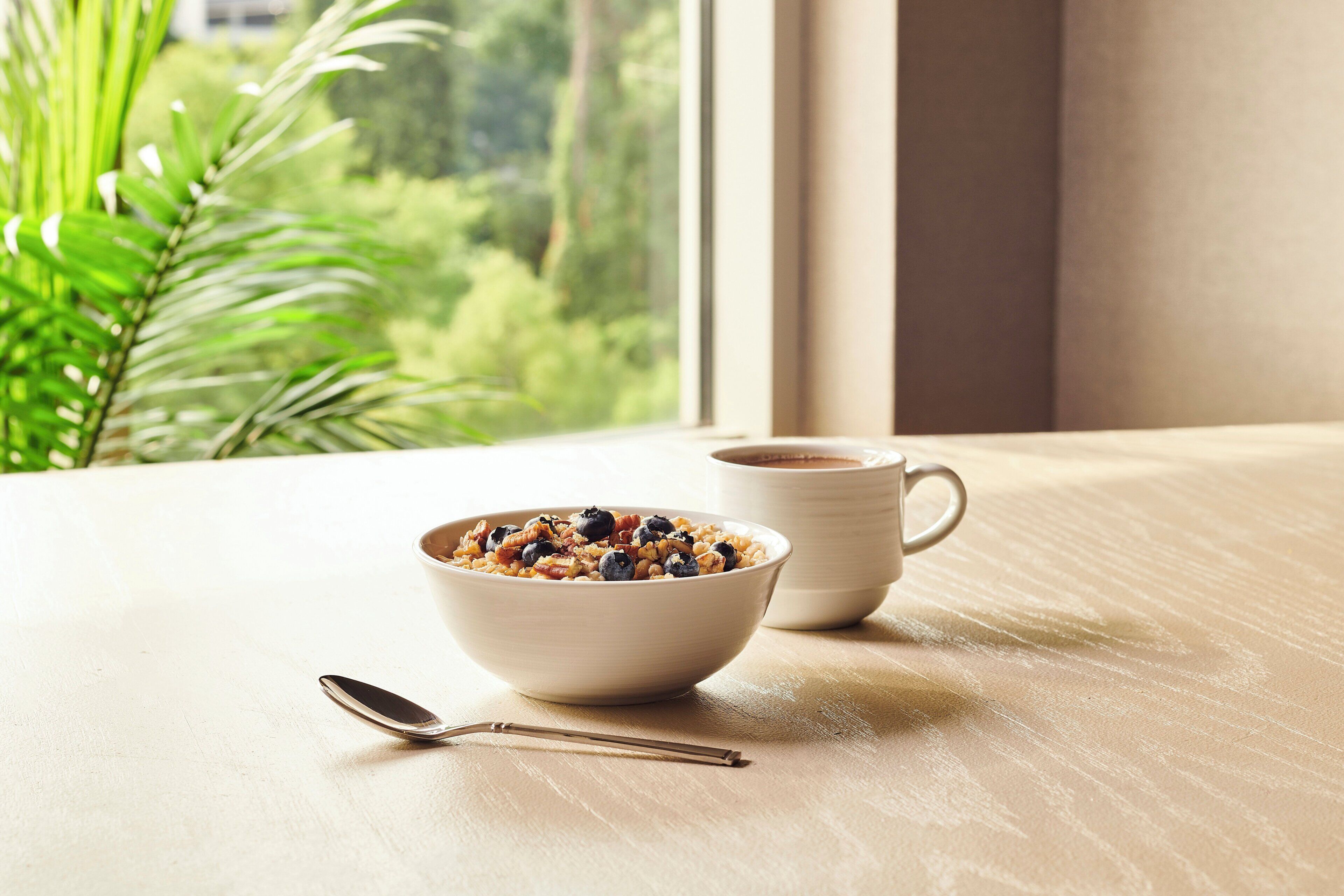 A bowl of cereal with a spoon on a wooden table by a window with a view of greenery.