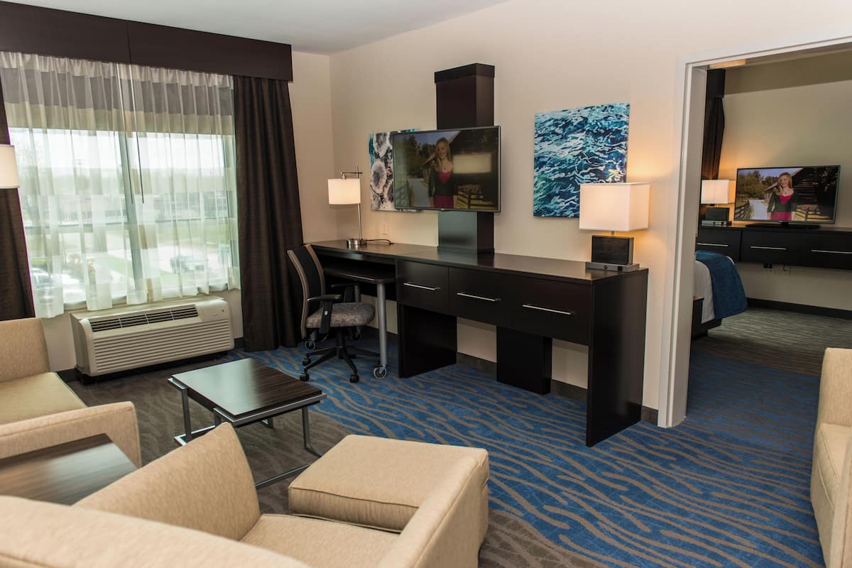 Habitación de hotel con escritorio, dos sillas, televisor y ventana grande con cortinas.