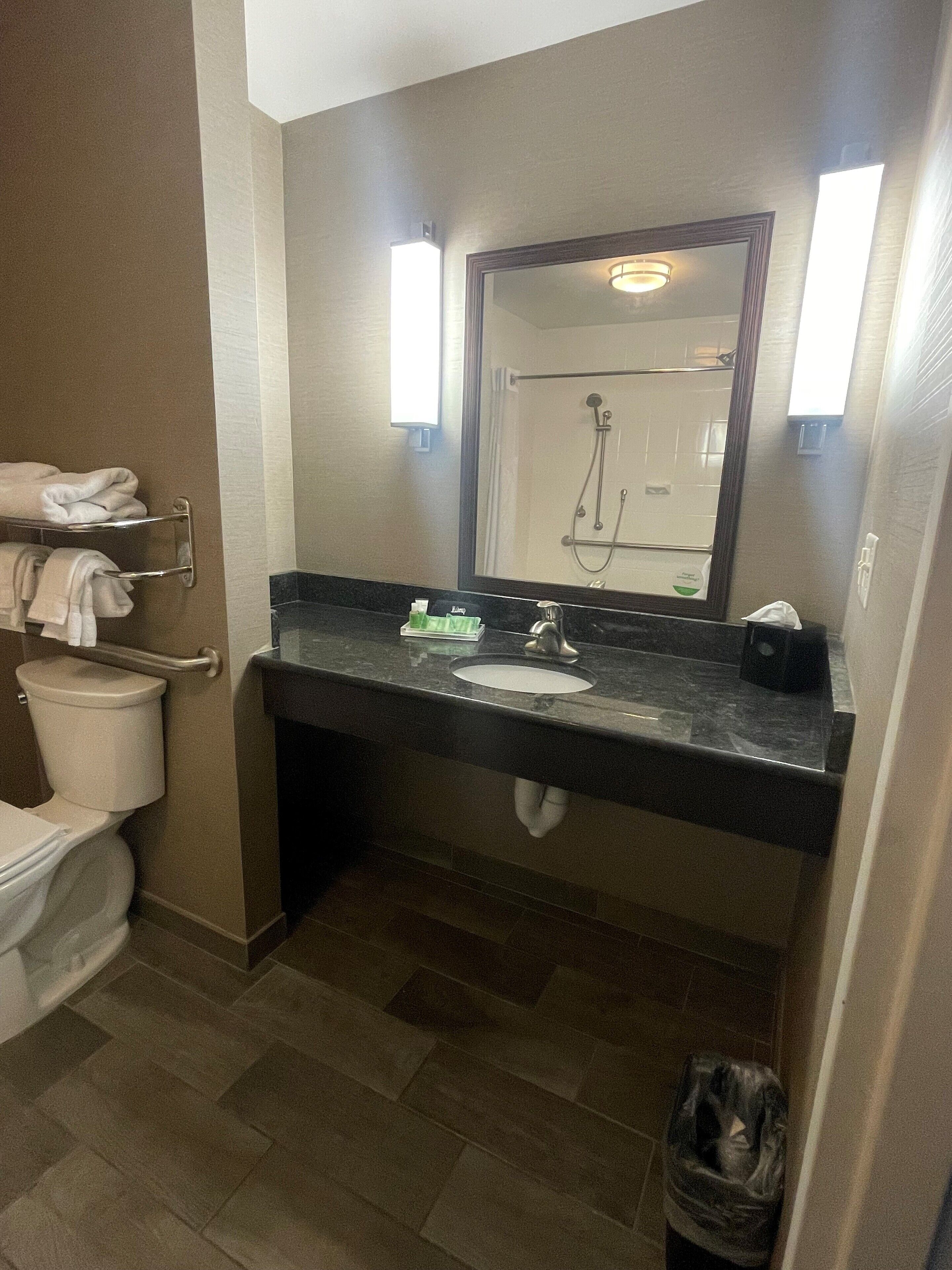 A bathroom with a toilet, sink, and mirror. There is a towel rack with folded towels, a trash can, and a soap dispenser on the sink.