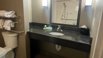 A bathroom with a toilet, sink, and mirror. There is a towel rack with folded towels, a trash can, and a soap dispenser on the sink.
