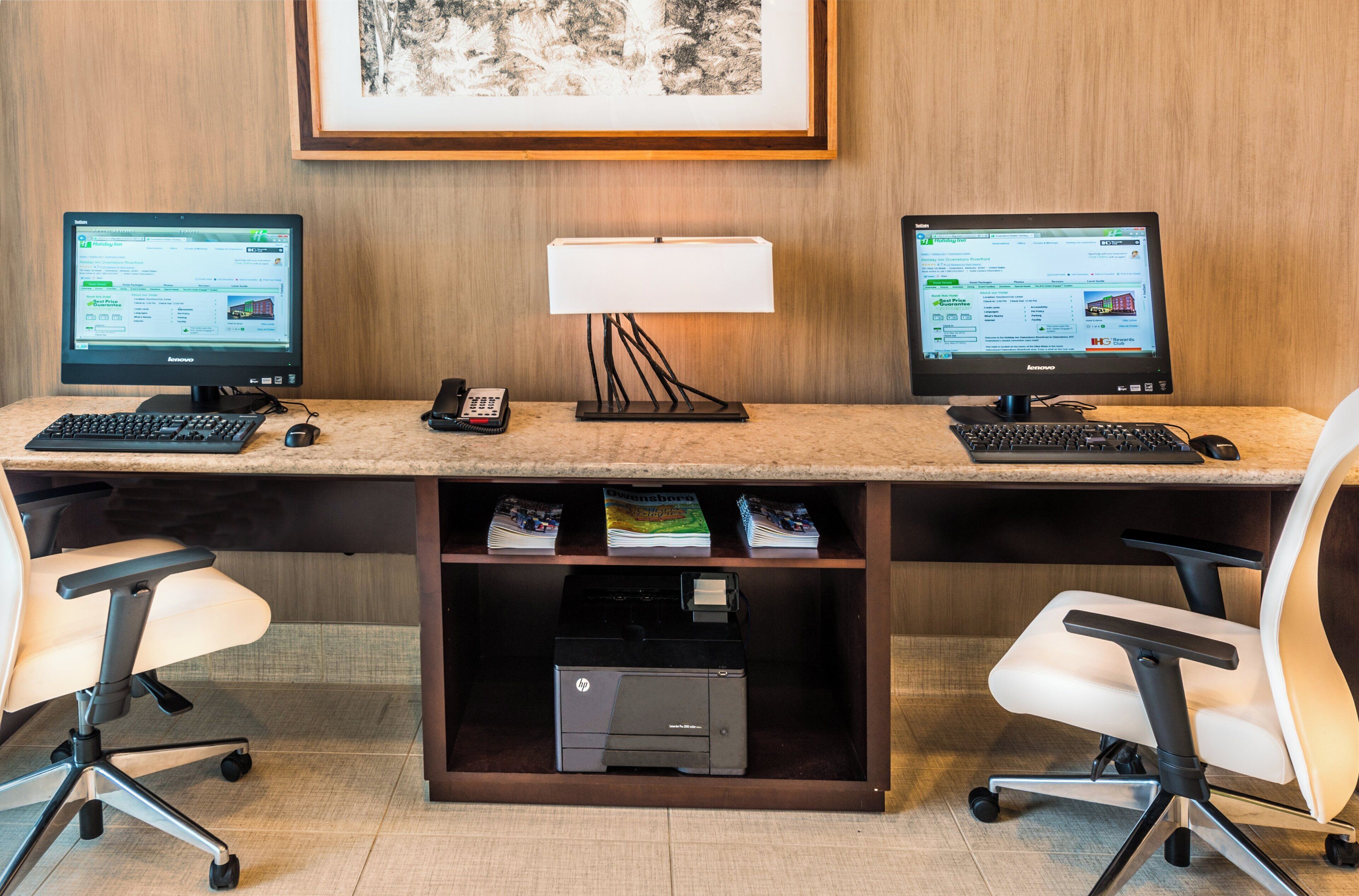 Un espacio de trabajo en un hotel con dos monitores, una lámpara de escritorio, un teléfono y una impresora HP sobre una encimera de mármol.