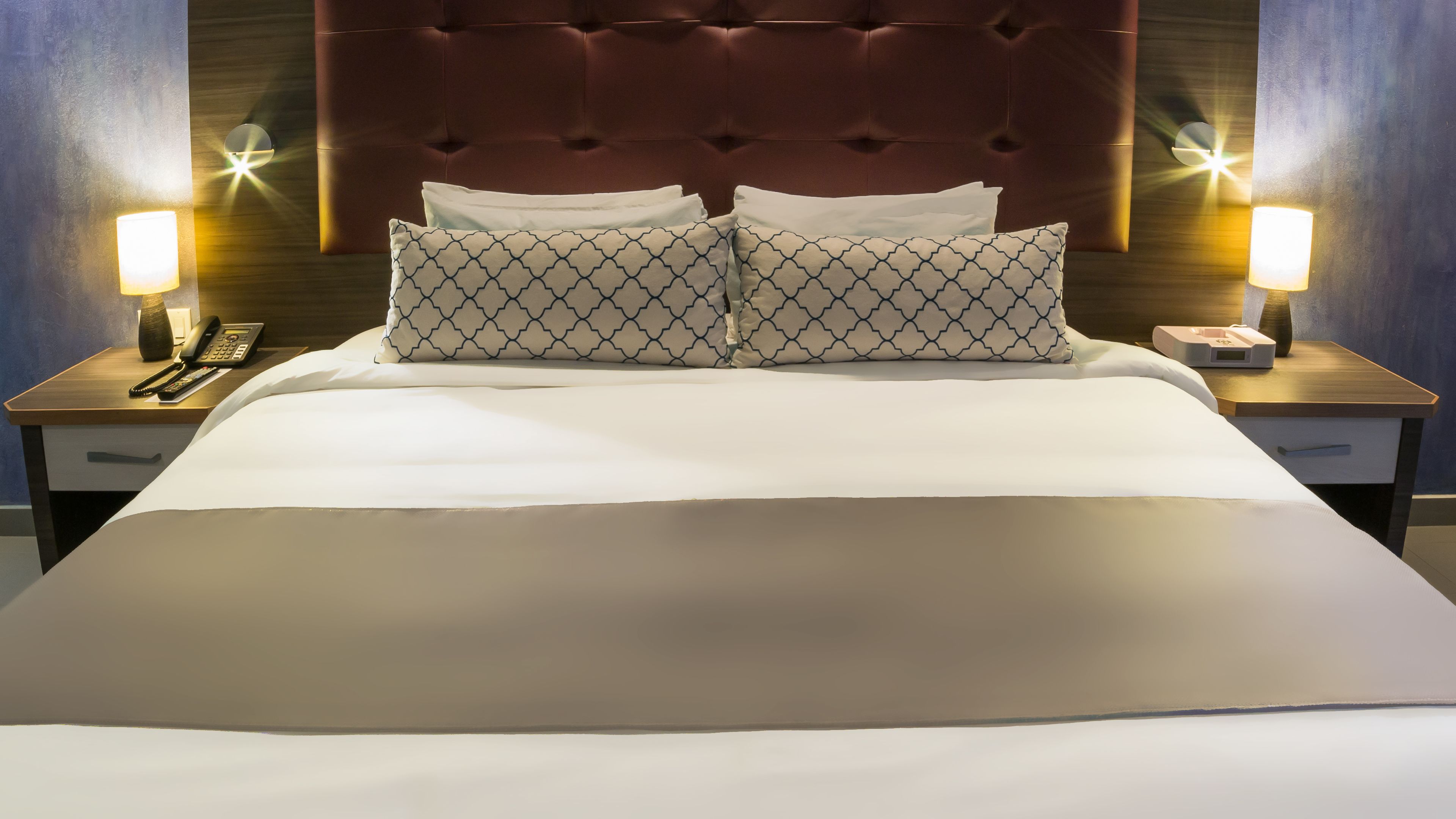 A neatly made bed with a large, padded headboard, two bedside tables with lamps, and a telephone on one of the tables.