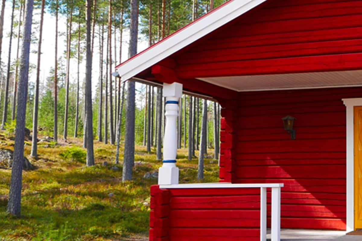 Una cabaña roja con puerta amarilla, barandilla de porche blanca y ventana con vista a un bosque.
