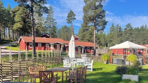 A campsite with wooden tables and chairs, a white umbrella, and red cabins in the background. - Hotell Moskogen (Leksand)