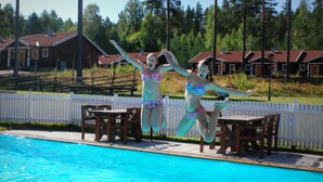 A swimming pool with two people jumping into it. There is a white picket fence and wooden chairs around the pool. - Hotell Moskogen (Leksand)