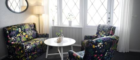 A room with a round white table, two floral-patterned armchairs, a round mirror, and a window with white curtains.