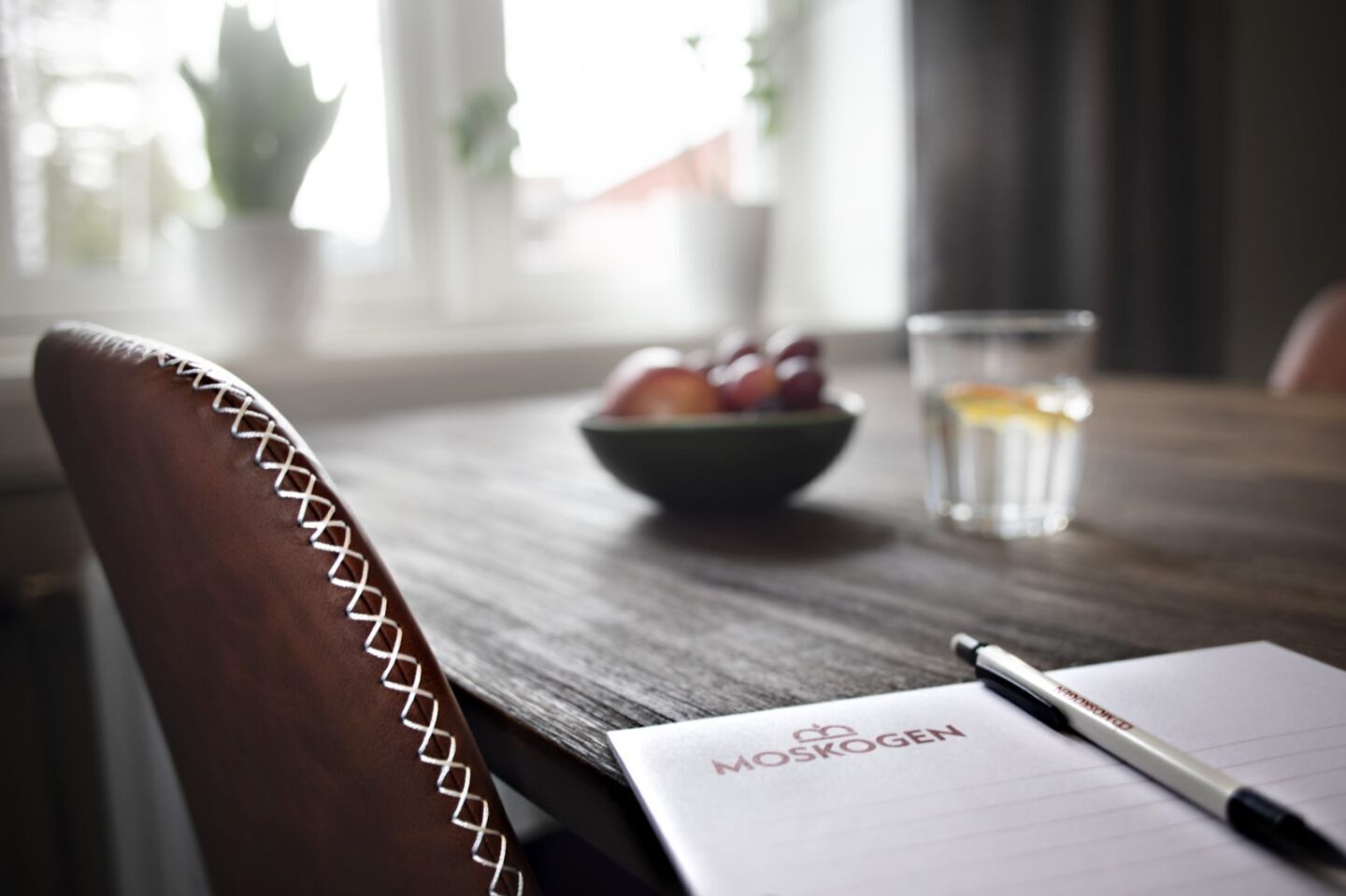 Una mesa de madera para comer con un bol de fruta y un vaso de agua. Hay un cuaderno y un bolígrafo sobre la mesa.