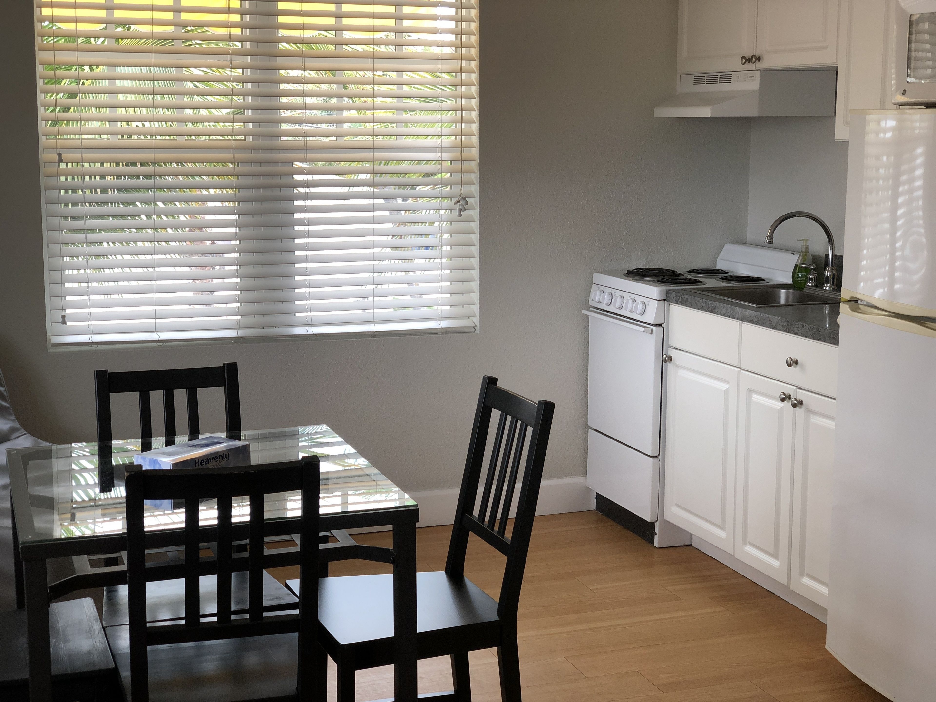 A compact kitchen with white cabinets, a stove, and a sink. There is a dining table with chairs and a window with blinds.