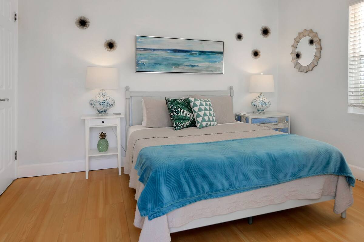 A bedroom with a bed, white nightstands, a painting, and a window with blinds.