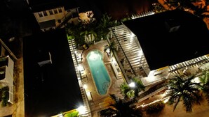 Vista aérea de un hotel con piscina rodeada de palmeras y una escalera que conduce a una terraza en la azotea.