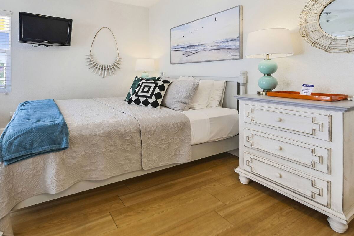 A bedroom with a bed, a dresser, a TV, a painting, and a window with blinds.