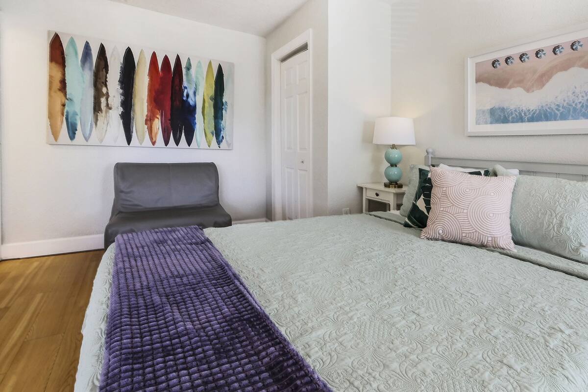 A neatly arranged bedroom with a bed, a chair, a nightstand, a lamp, and a wall art piece.