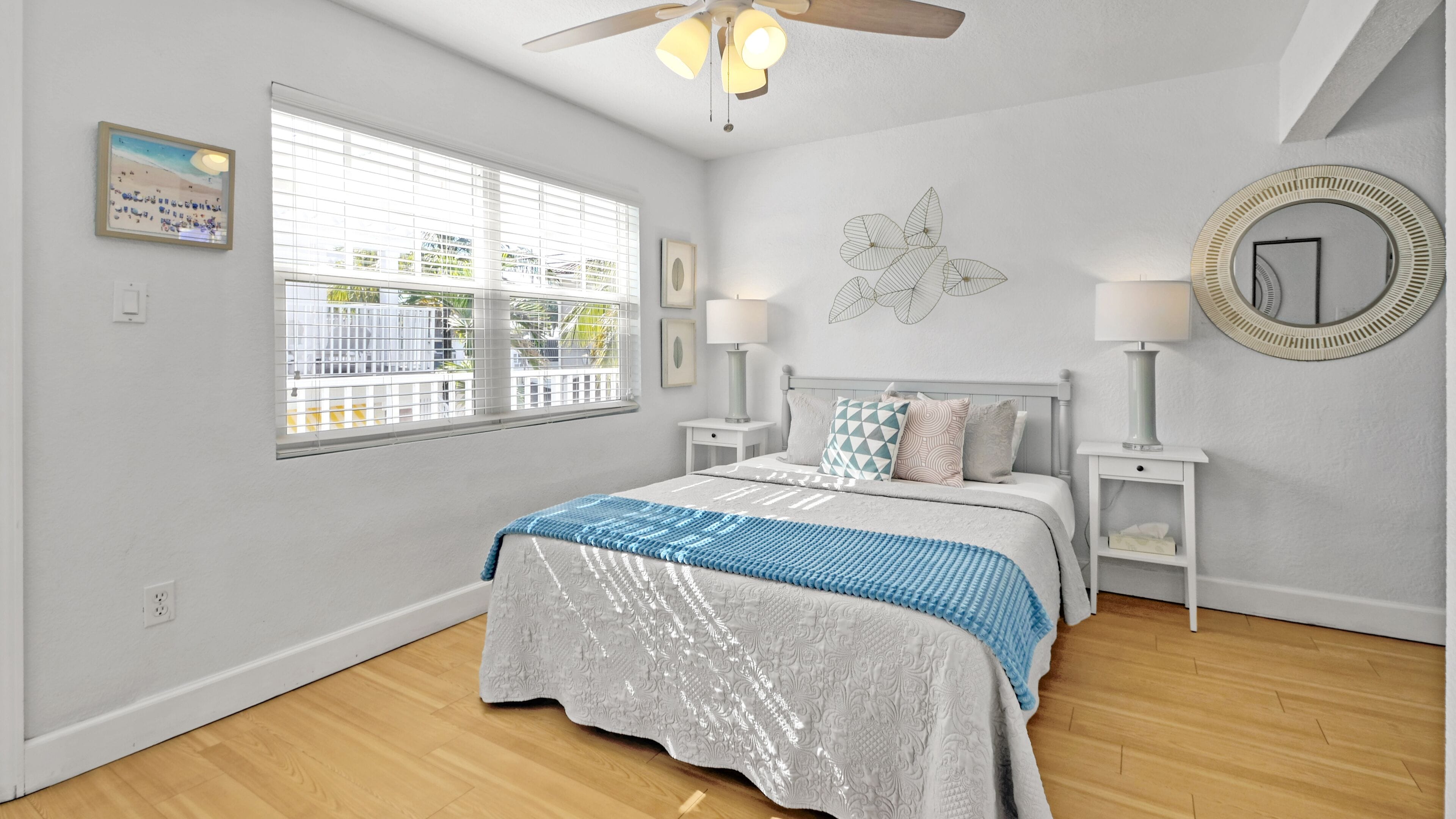 A bedroom with a bed, two bedside tables, a ceiling fan, a window with blinds, and a wall-mounted mirror.