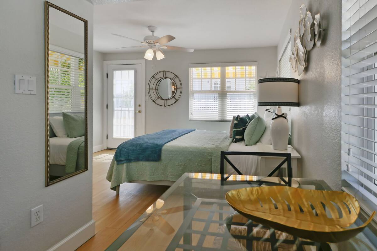 A bedroom with a bed, a desk, a chair, a ceiling fan, a window with blinds, and a mirror.