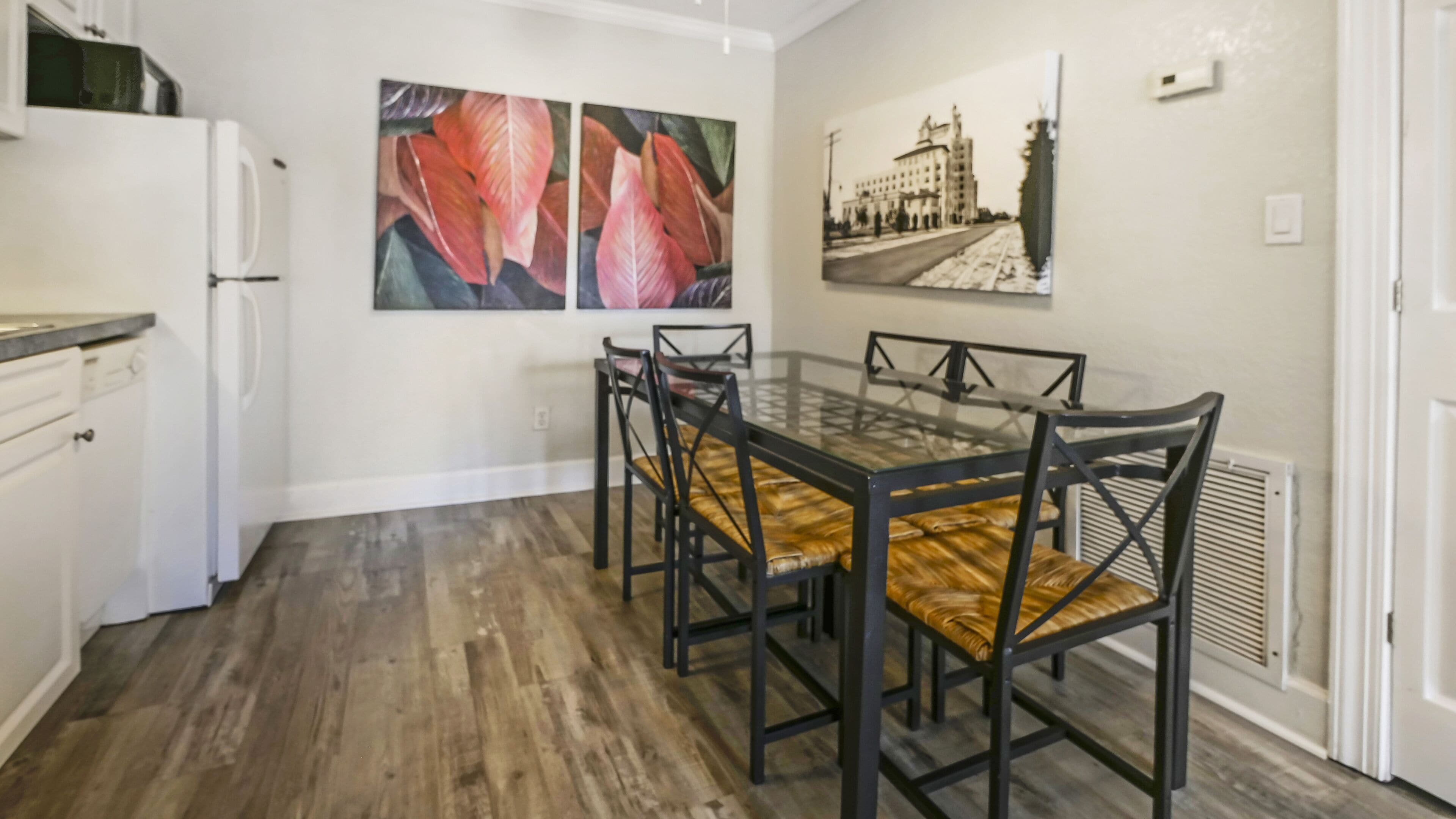 A dining area with a glass table and chairs, a white refrigerator, and two paintings on the wall.