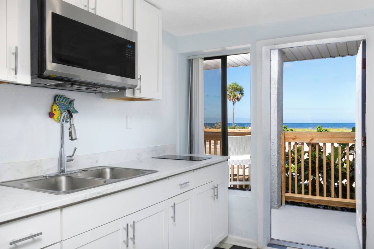 Una cocina con microondas, pileta doble y vista a la playa a través de una puerta abierta.