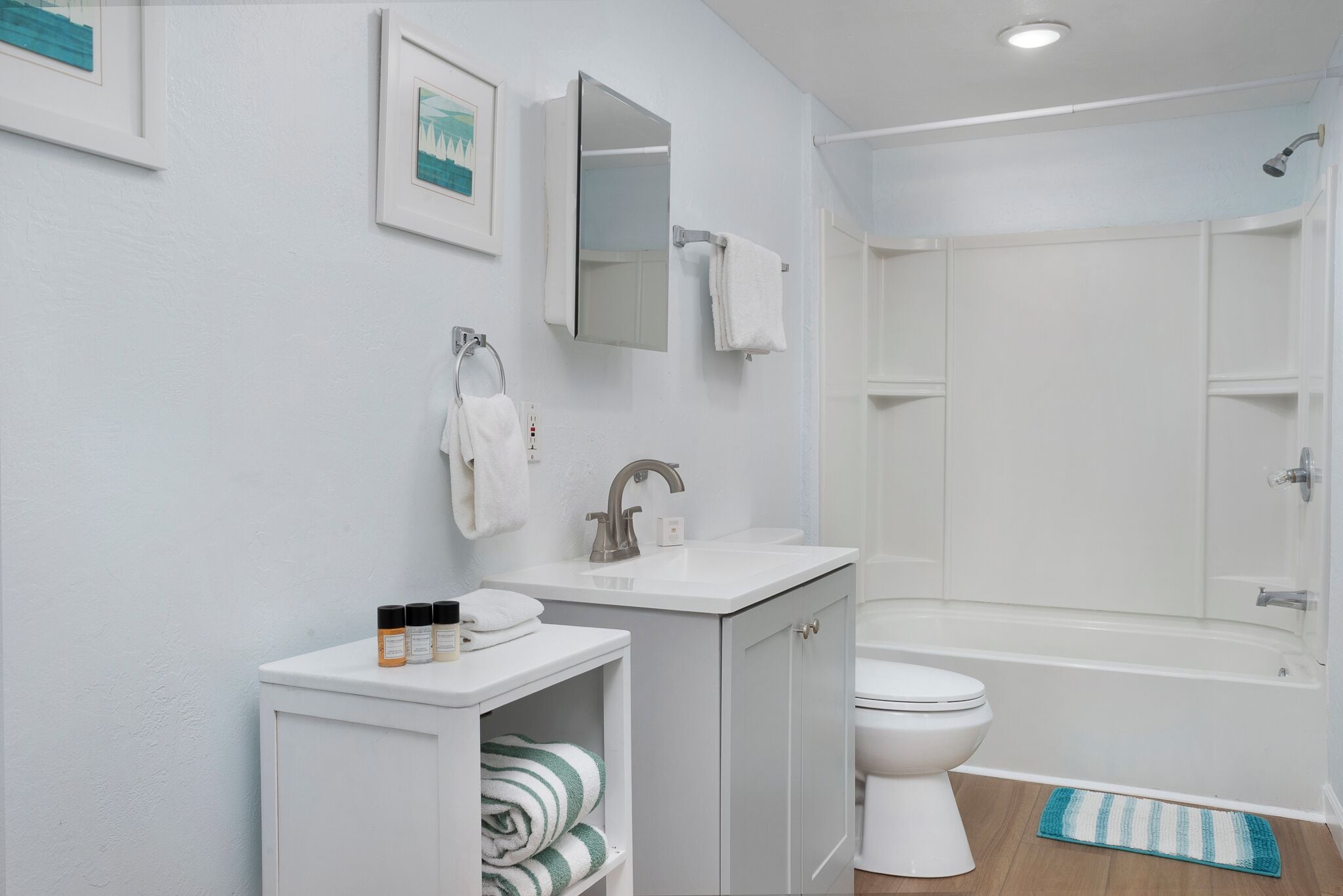 A modern bathroom with a white sink, vanity, and toilet. There is a shower area with a glass door and a blue mat.