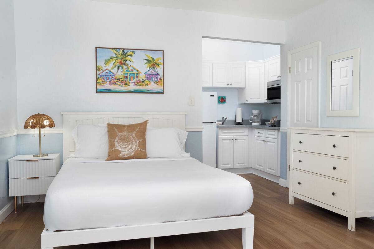 Un dormitorio con cama, mesita de noche, lámpara y un cuadro de casas tropicales.