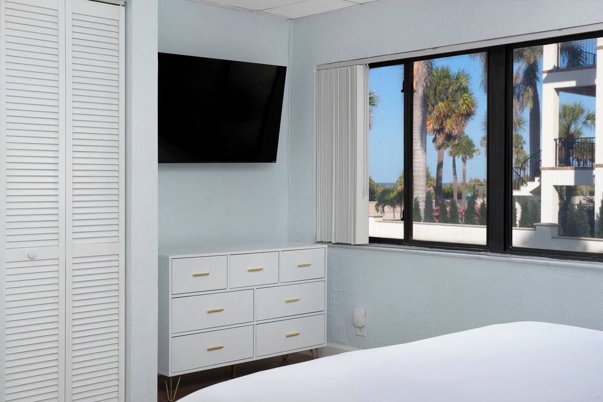 Un dormitorio con cama, televisor montado en la pared, ventana con vista a palmeras y cómoda blanca con cajones.