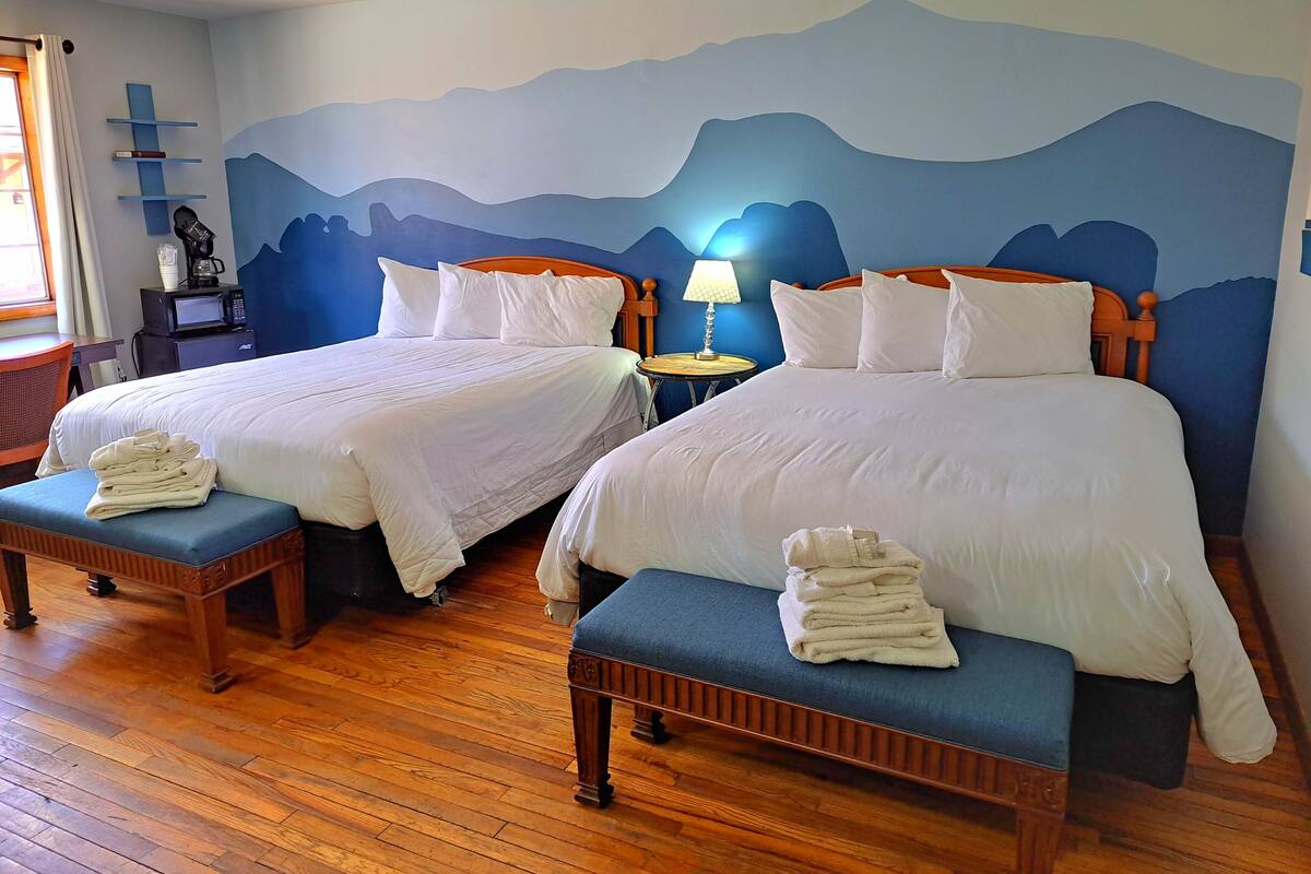 A hotel room with two beds, wooden headboards, a wooden floor, a desk with a chair, a lamp, and a wall with a mountain mural.