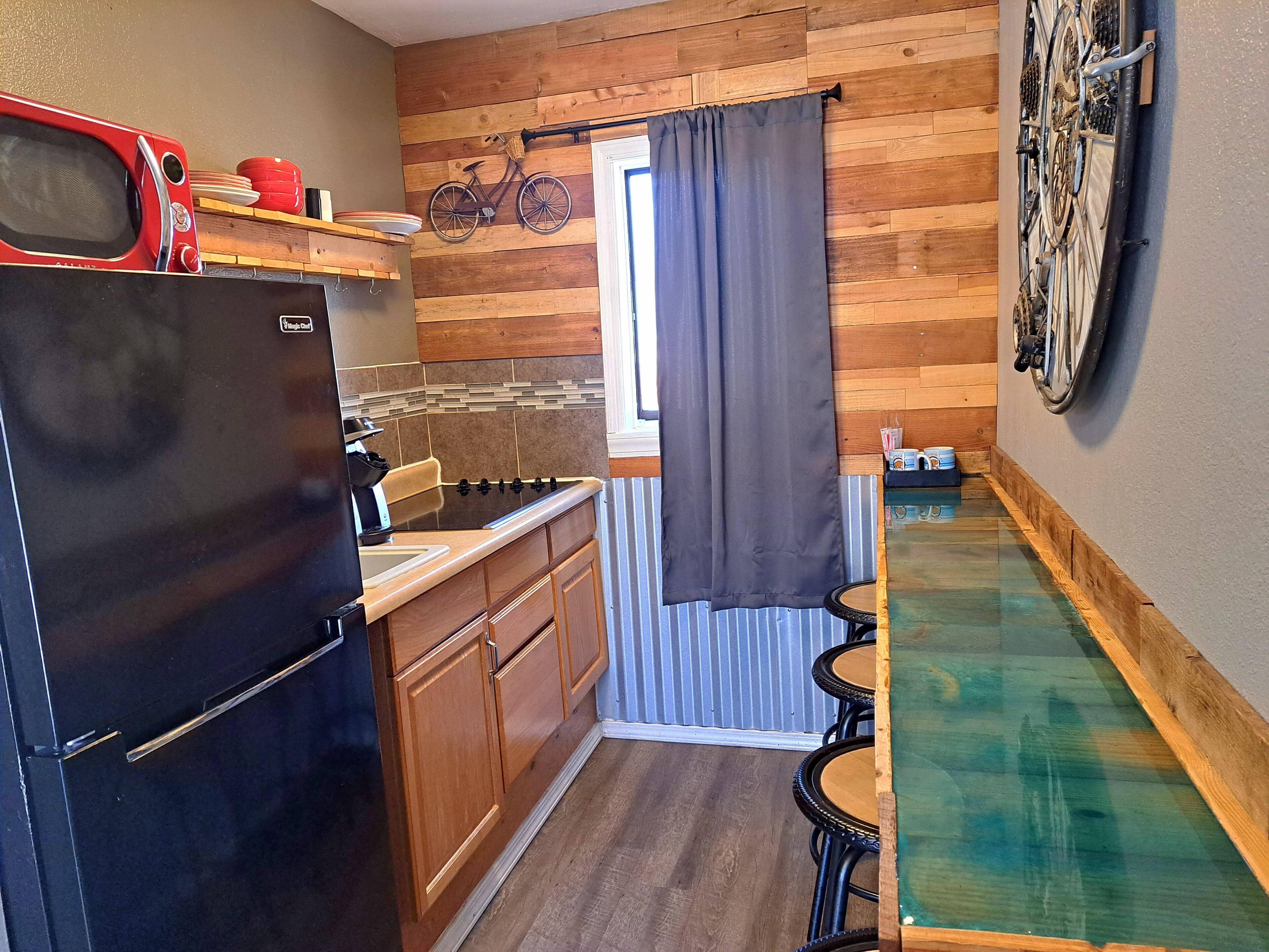 Une cuisine compacte avec un revêtement mural en bois, un réfrigérateur, un four à micro-ondes et un petit coin repas comprenant une table et des chaises en verre.