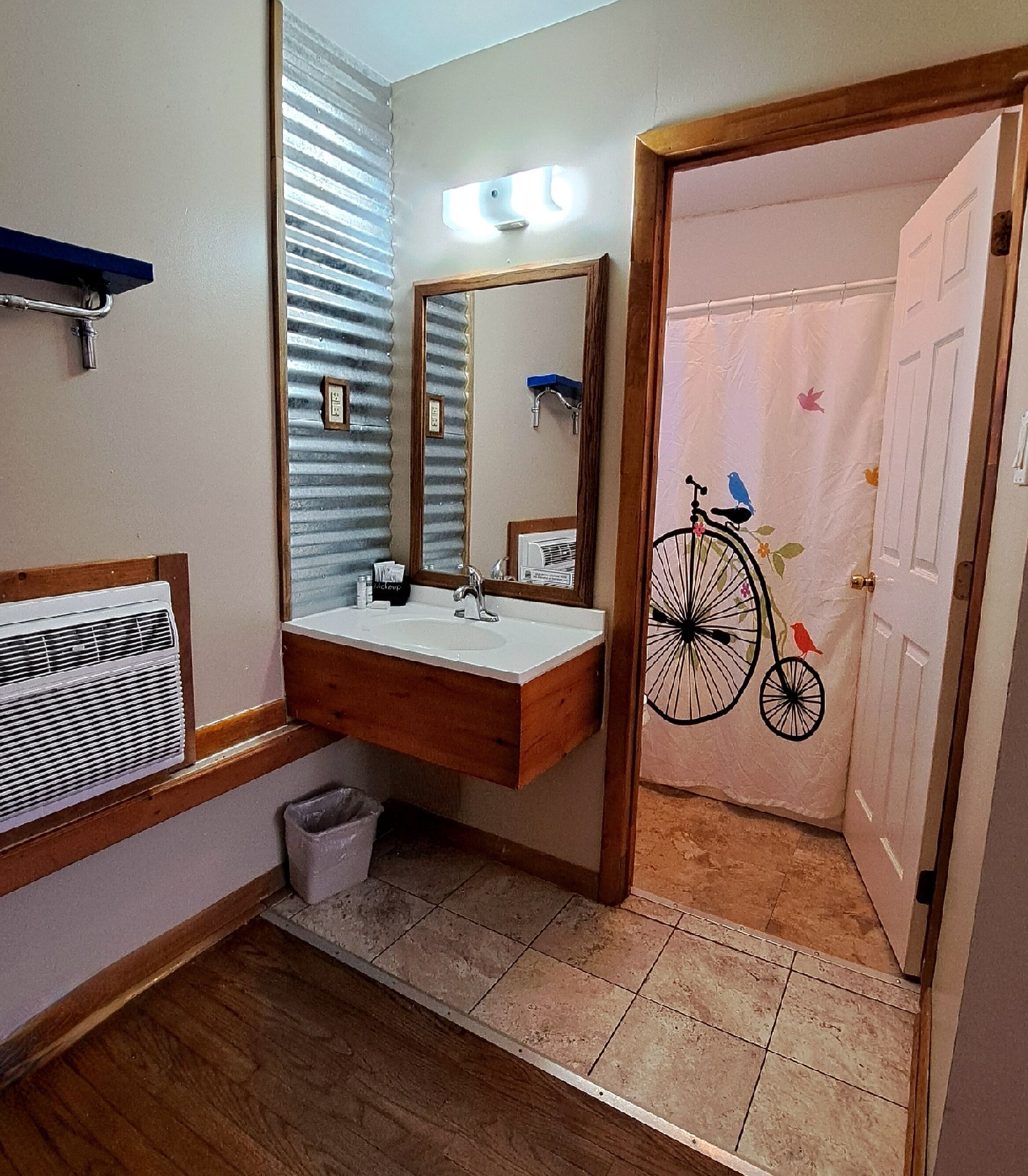 Une salle de bain avec un meuble-lavabo en bois, un miroir, un évier, une fenêtre avec des stores, un rideau de douche orné d’un motif de vélo et une porte.