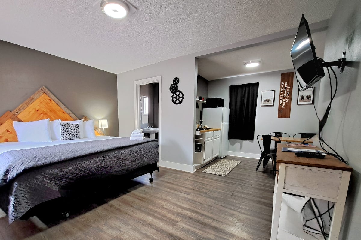 A hotel room with a bed, a desk, a chair, a TV, and a kitchenette.