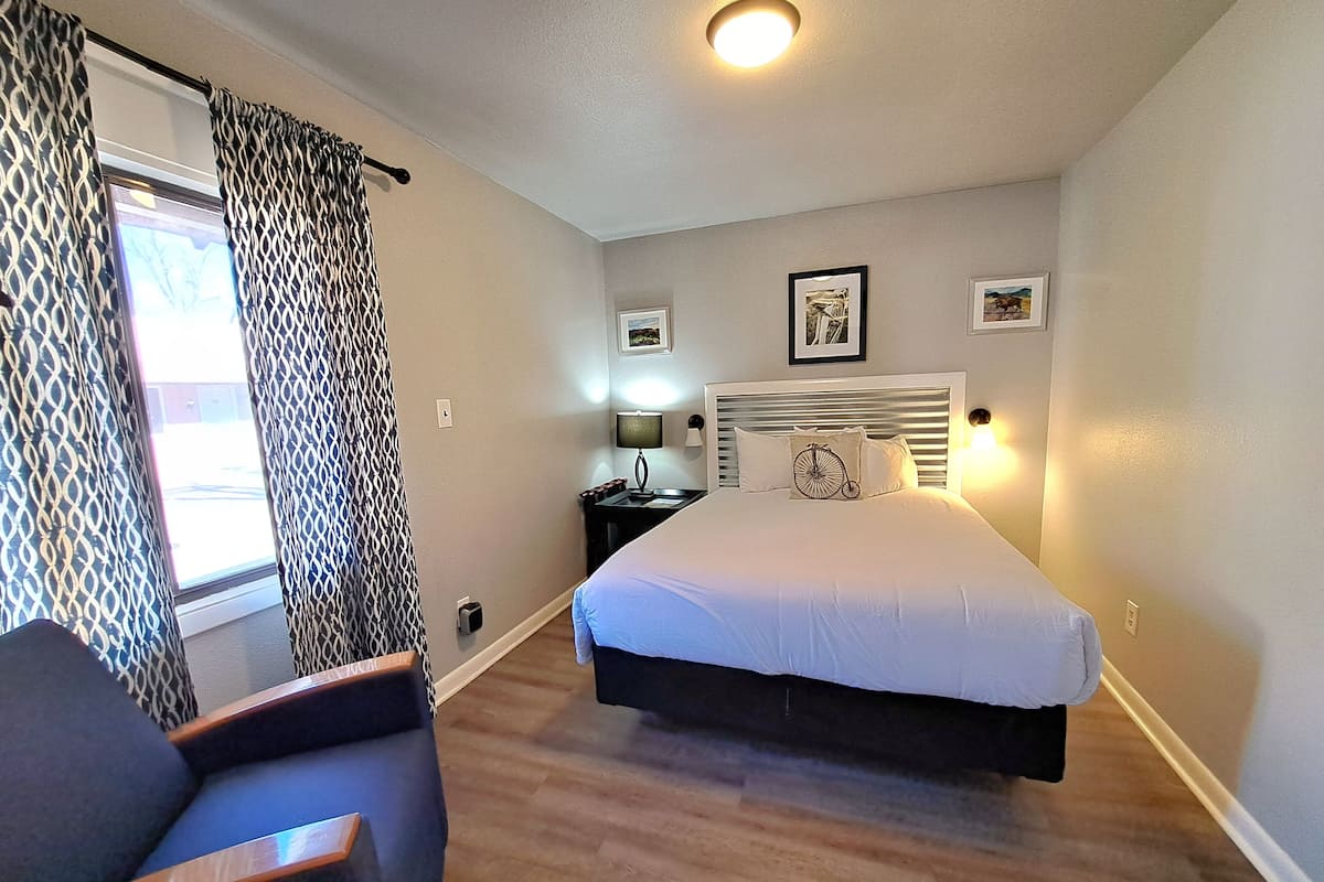 A bedroom with a bed, a chair, a nightstand, and a window with patterned curtains.