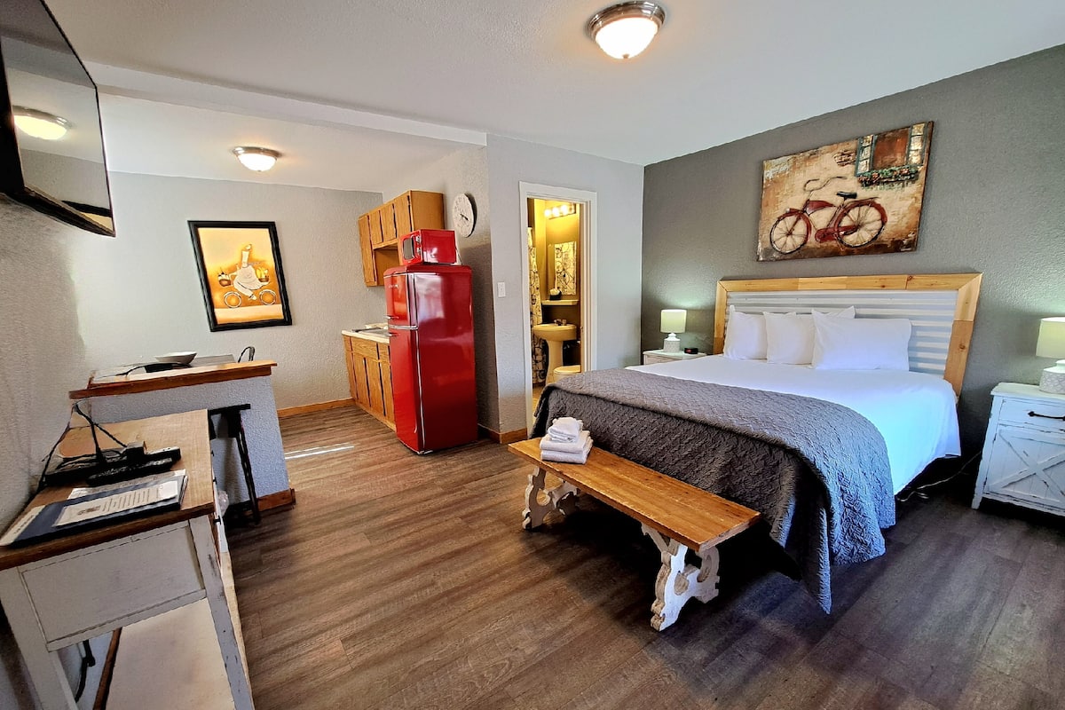 A compact hotel room with a bed, bedside tables, a small desk, a red refrigerator, a wooden bench, and a painting of a bicycle.