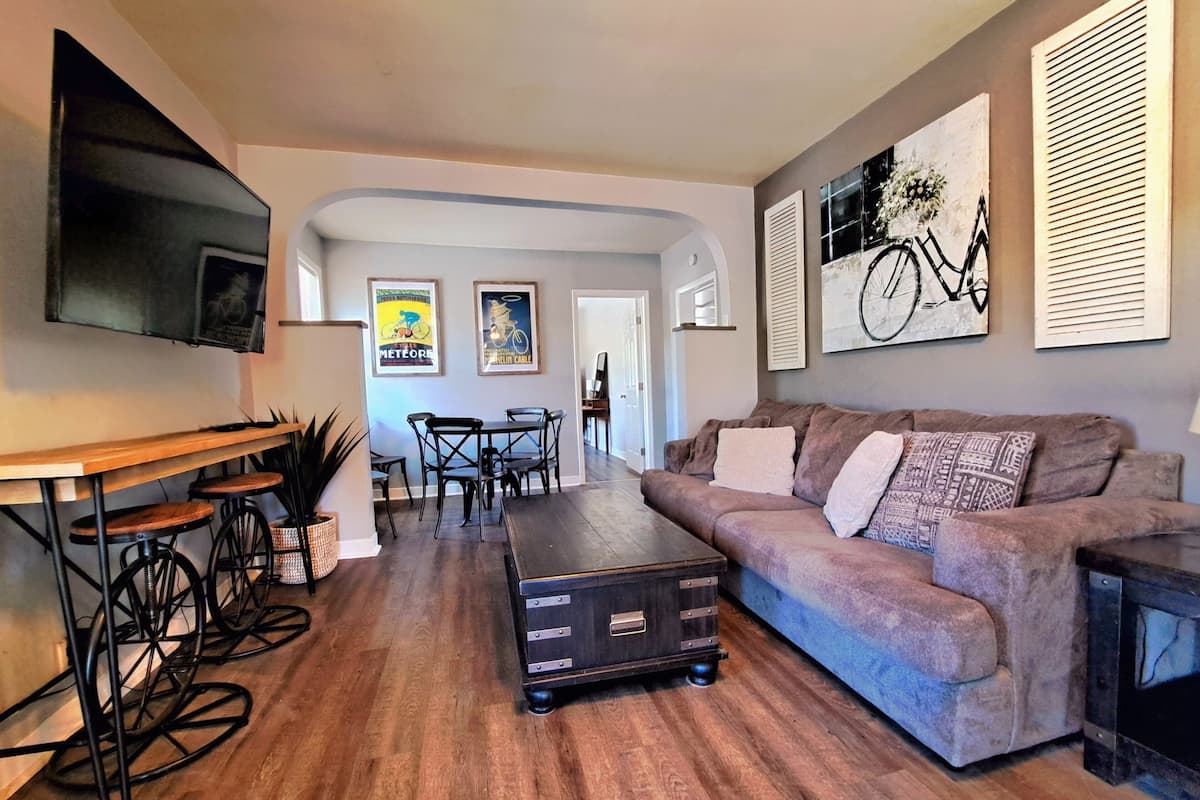 A modern living room with a sofa, a coffee table, and a TV. There is a dining area with chairs and a table.