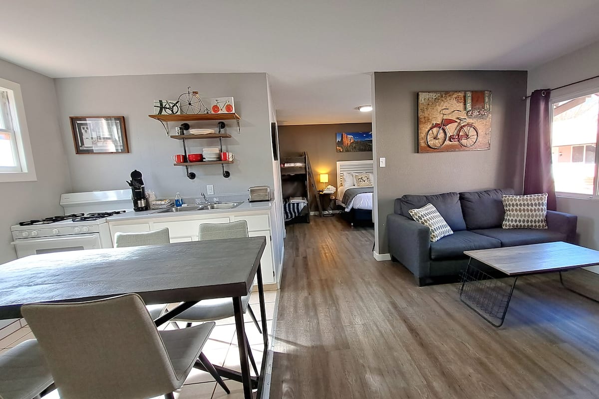 A modern kitchen with white appliances, a dining area with a table and chairs, a gray sofa with patterned cushions, and a large window with curtains.