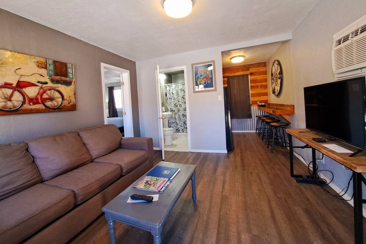 A living room with a brown sofa, a coffee table with magazines, a TV, and a wooden dining table with chairs.