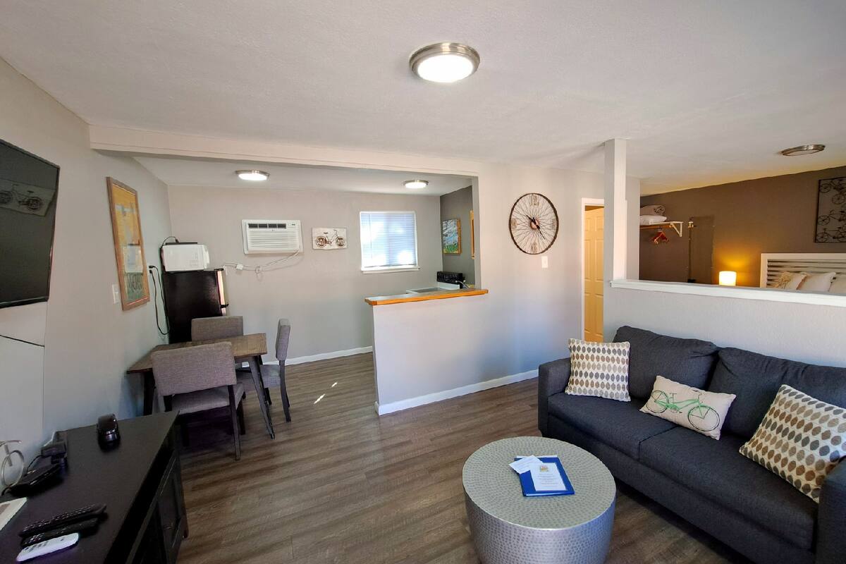 A living room with a sofa, a coffee table, a dining area with a table and chairs, a kitchen area with a counter, and a small window.
