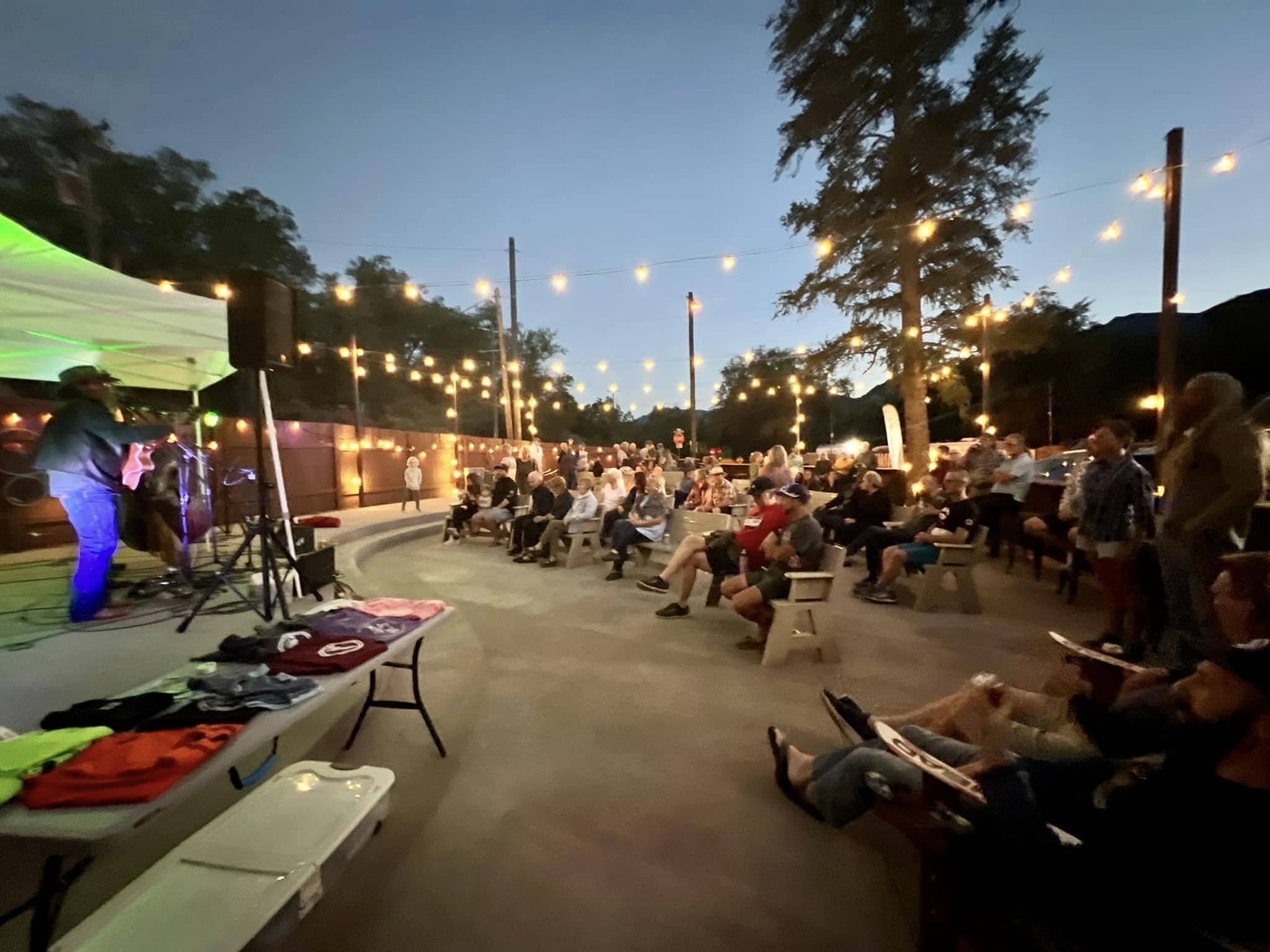 An outdoor event with a stage and seated audience, surrounded by trees and string lights.